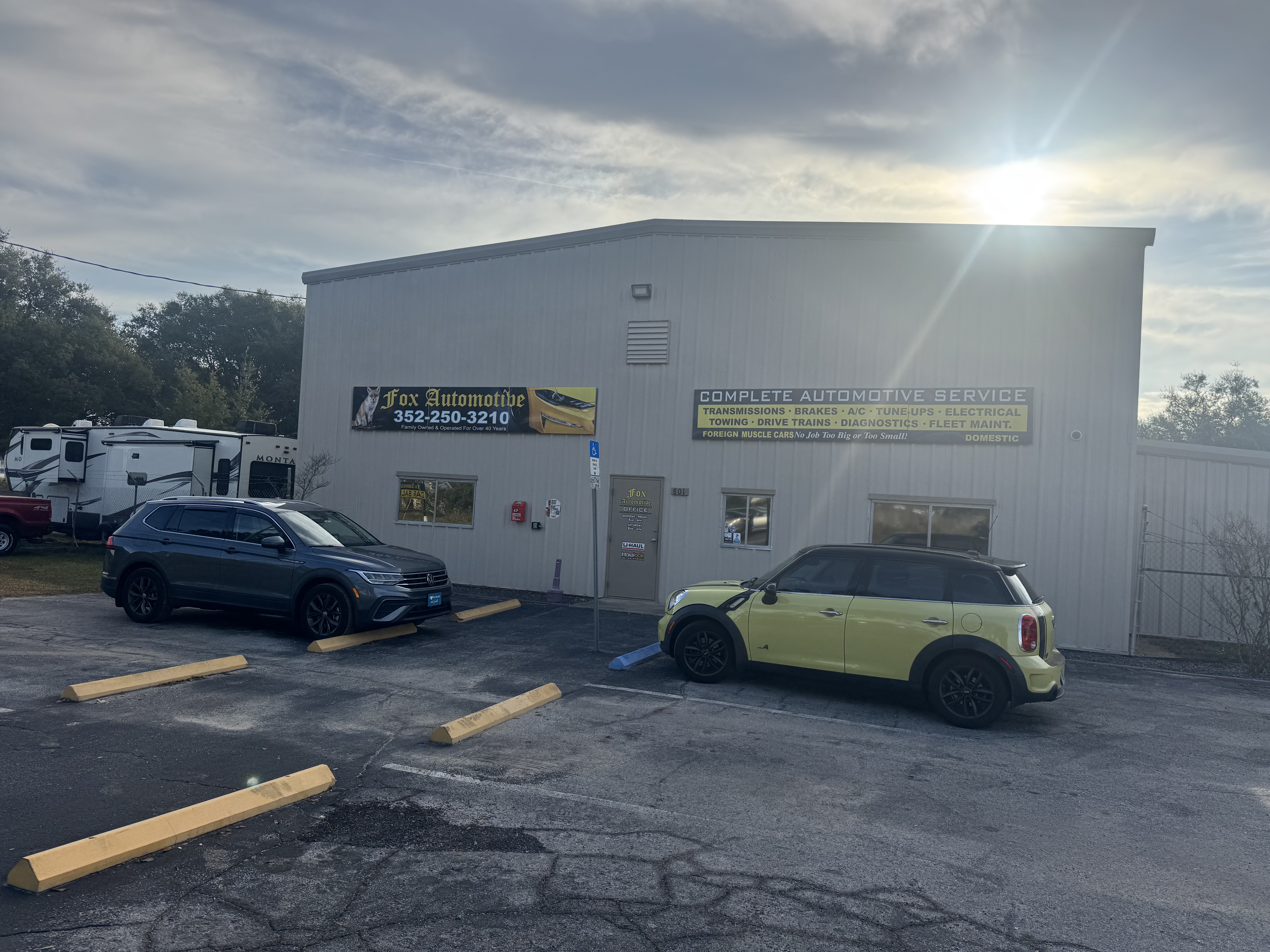 Exterior view of Fox Automotive building with white walls, blue and yellow signage displaying phone number 352-250-3210, and parked vehicles including a dark SUV and yellow Mini Cooper in front