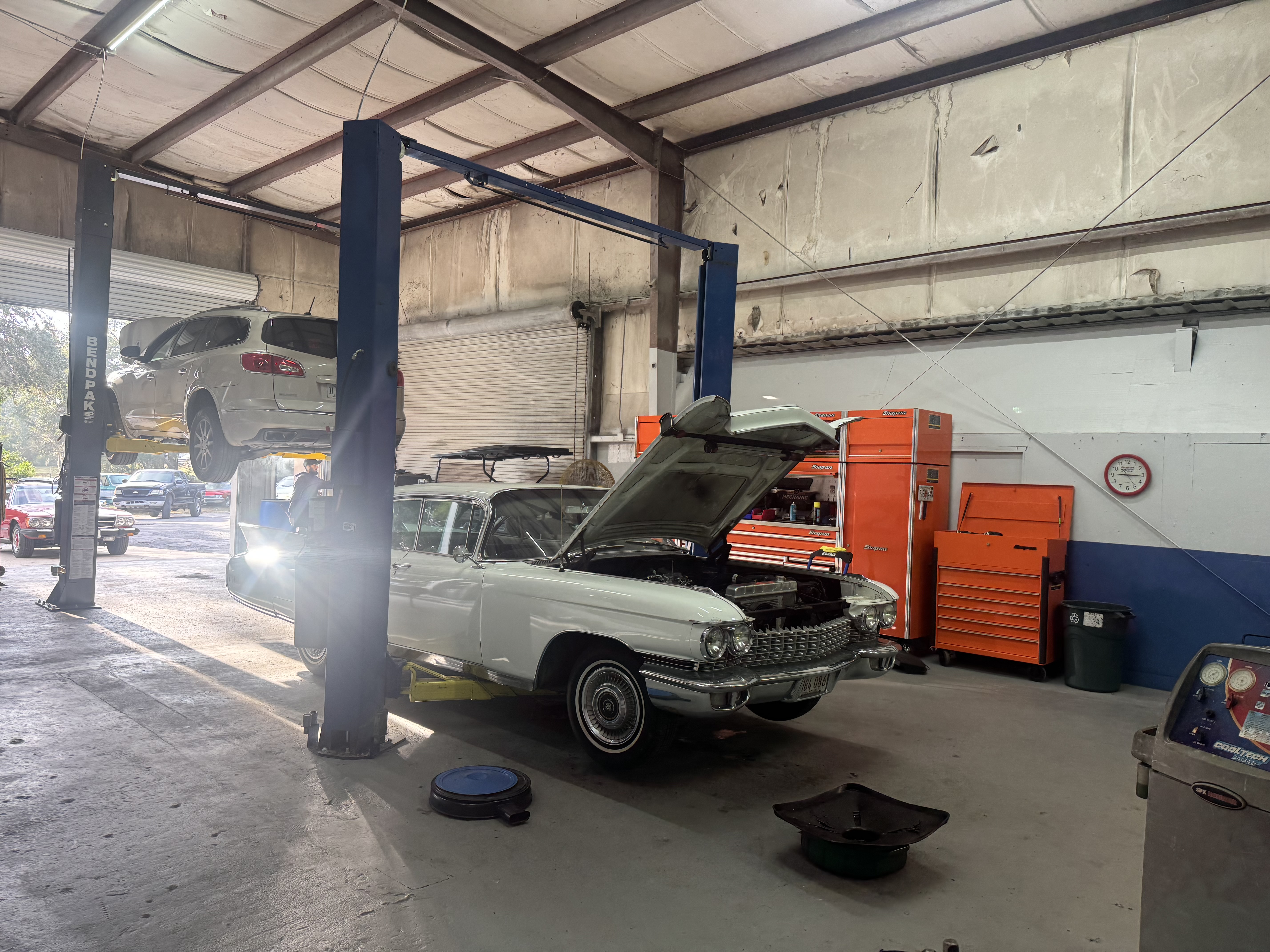 Interior of Fox Automotive garage bay showing a white vintage Cadillac with hood open being serviced, with a modern SUV on a lift in the background and orange tool chests along the wall