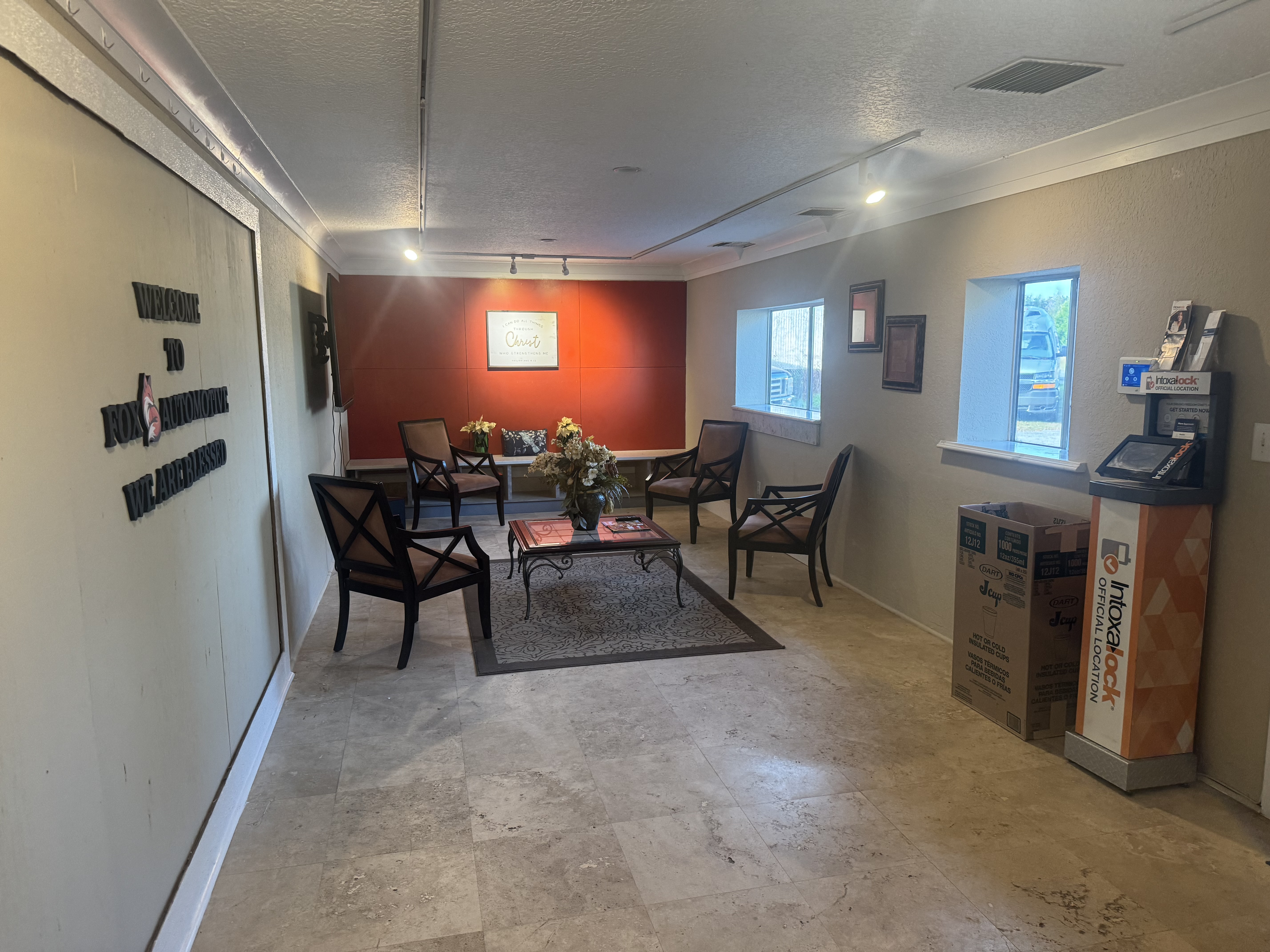 Customer waiting area at Fox Automotive featuring comfortable seating arrangements with dark wood chairs, area rugs, red accent wall, and NICOlock payment kiosk