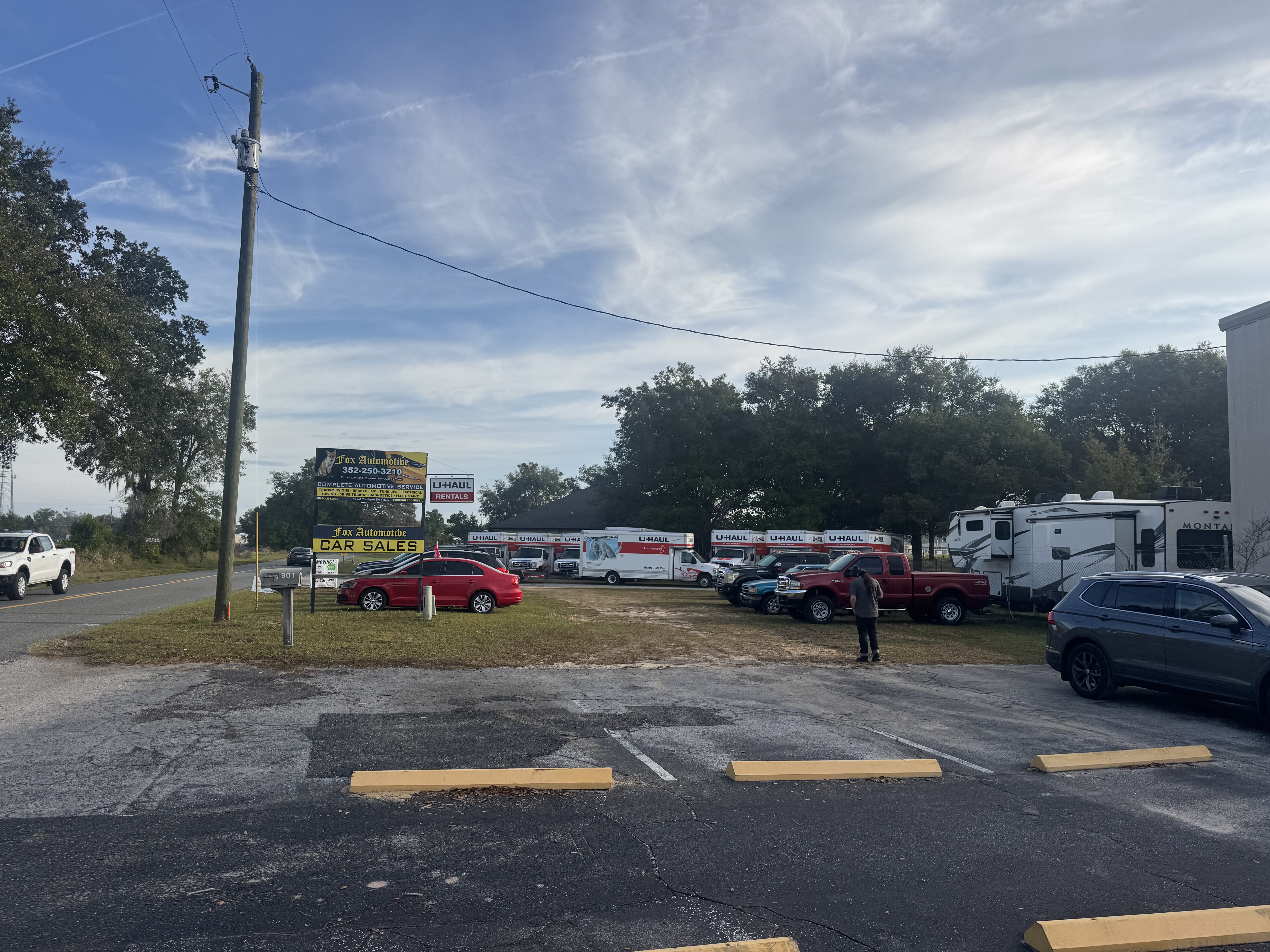 Parking lot view of Fox Automotive showing U-Haul rental trucks and vehicles, with business signage visible and trees in the background